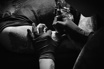 A close-up of a tattoo artist at work, using a tattoo machine on a client's skin with visible tattoos on the arm. The artist is wearing black gloves and the lighting creates a dramatic contrast.