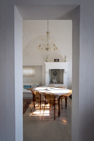 A clean, minimalist room with light-colored walls and a high arched ceiling. A wooden dining table with a lace tablecloth and six transparent chairs occupies the foreground. There is a white fireplace in the background, flanked by small statues and framed pictures. A chandelier hanging from the ceiling contributes to the elegant ambiance. Sunlight streams in, casting shadows on the floor.
