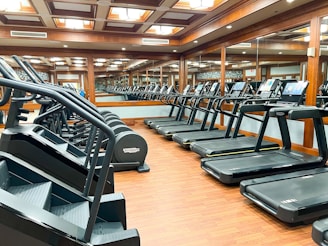 A modern gym outfitted with rows of treadmills and step machines, organized neatly in a spacious room with wooden floors. Large mirrors line the walls, enhancing the perception of space and reflecting the equipment.