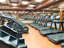 A modern gym outfitted with rows of treadmills and step machines, organized neatly in a spacious room with wooden floors. Large mirrors line the walls, enhancing the perception of space and reflecting the equipment.