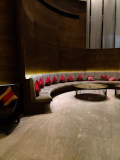 A modern seating area with a curved, cushioned sofa adorned with numerous red cushions. The background features a combination of textured wooden and metallic surfaces, while two round, metal-framed tables occupy the center. Soft lighting creates a warm ambiance, highlighting the sleek design and the polished marble floor.