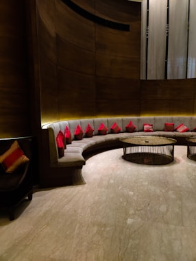 A modern seating area with a curved, cushioned sofa adorned with numerous red cushions. The background features a combination of textured wooden and metallic surfaces, while two round, metal-framed tables occupy the center. Soft lighting creates a warm ambiance, highlighting the sleek design and the polished marble floor.