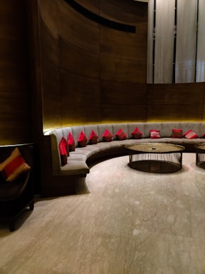 A modern seating area with a curved, cushioned sofa adorned with numerous red cushions. The background features a combination of textured wooden and metallic surfaces, while two round, metal-framed tables occupy the center. Soft lighting creates a warm ambiance, highlighting the sleek design and the polished marble floor.
