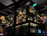A dimly lit restaurant interior with modern industrial design. Several pendant lamps with warm lighting hang from the ceiling. The back wall features a large shelving unit with potted plants and red containers. A green exit sign is visible above. Chalkboard-style artwork decorates the walls with culinary themes.