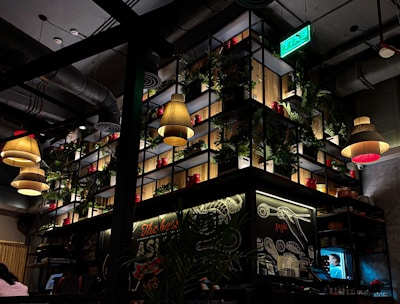 A dimly lit restaurant interior with modern industrial design. Several pendant lamps with warm lighting hang from the ceiling. The back wall features a large shelving unit with potted plants and red containers. A green exit sign is visible above. Chalkboard-style artwork decorates the walls with culinary themes.