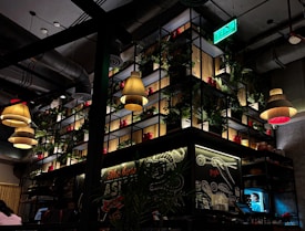 A dimly lit restaurant interior with modern industrial design. Several pendant lamps with warm lighting hang from the ceiling. The back wall features a large shelving unit with potted plants and red containers. A green exit sign is visible above. Chalkboard-style artwork decorates the walls with culinary themes.