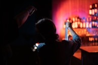 A dimly lit scene featuring two silhouetted individuals, with one person holding a strip of film in their hands. In the background, shelves are lined with illuminated bottles of various shapes, casting a warm, colorful glow.
