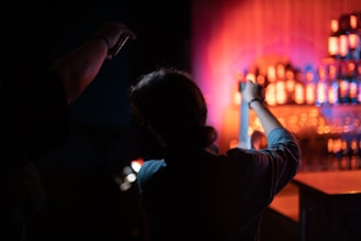 A dimly lit scene featuring two silhouetted individuals, with one person holding a strip of film in their hands. In the background, shelves are lined with illuminated bottles of various shapes, casting a warm, colorful glow.