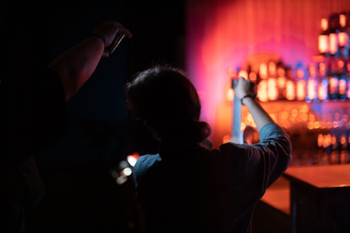 A dimly lit scene featuring two silhouetted individuals, with one person holding a strip of film in their hands. In the background, shelves are lined with illuminated bottles of various shapes, casting a warm, colorful glow.