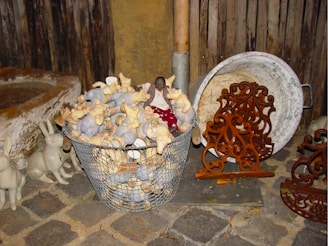 A collection of plush toy pigs fills a metal basket on a cobblestone surface, accompanied by ornamental metalwork and rabbit decorations nearby. The rustic setting includes a weathered wooden backdrop and a large, round metal container.