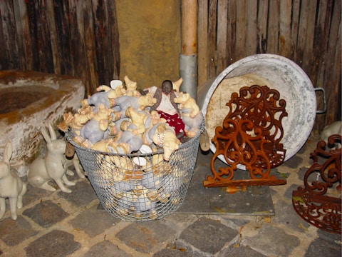 A collection of plush toy pigs fills a metal basket on a cobblestone surface, accompanied by ornamental metalwork and rabbit decorations nearby. The rustic setting includes a weathered wooden backdrop and a large, round metal container.
