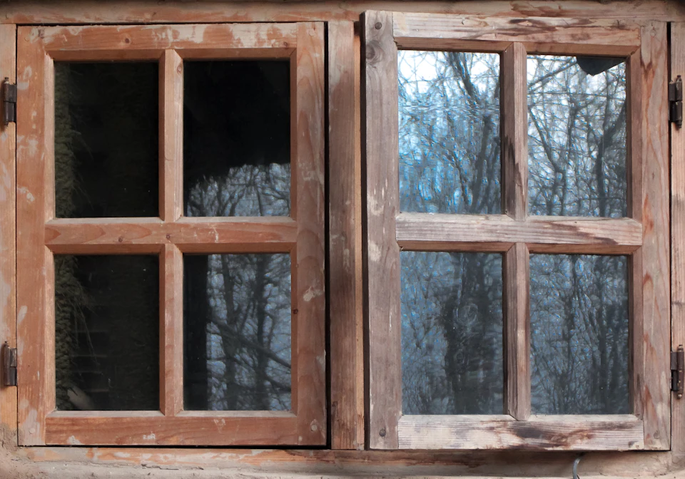 Old wooden windows with damaged right pane partially open, showing tree-lined landscape outside