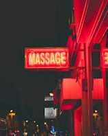 A brightly lit red neon sign with the word 'MASSAGE' hangs on the side of a building. The sign is illuminated against a dark urban background with other lights and signs in the distance. The image appears to be taken at night in an urban setting with a bustling street scene.