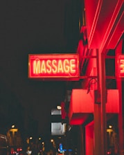 A brightly lit red neon sign with the word 'MASSAGE' hangs on the side of a building. The sign is illuminated against a dark urban background with other lights and signs in the distance. The image appears to be taken at night in an urban setting with a bustling street scene.