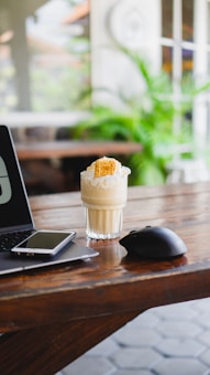 A glass of creamy iced coffee topped with whipped cream and a wafer sits on a wooden table. Beside it are a smartphone and a closed laptop, indicating a workspace setting. A computer mouse rests next to the glass, and there are blurred green plants in the background, suggesting an indoor or semi-outdoor environment.