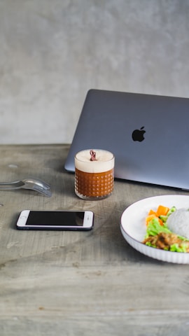 A setting on a wooden table featuring a smartphone, a glass with a creamy beverage topped with a garnish, a sleek laptop with a visible logo, and a plate of food including rice, vegetables, and a fork placed beside the phone.