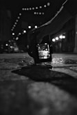 A black and white photograph showing a hand holding a digital camera close to the ground. The camera screen displays a bright street scene with overhead string lights, while the background is blurred. The scene is captured in a dimly lit urban environment.