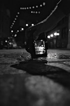 A black and white photograph showing a hand holding a digital camera close to the ground. The camera screen displays a bright street scene with overhead string lights, while the background is blurred. The scene is captured in a dimly lit urban environment.