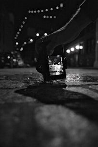 A black and white photograph showing a hand holding a digital camera close to the ground. The camera screen displays a bright street scene with overhead string lights, while the background is blurred. The scene is captured in a dimly lit urban environment.