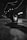 A black and white photograph showing a hand holding a digital camera close to the ground. The camera screen displays a bright street scene with overhead string lights, while the background is blurred. The scene is captured in a dimly lit urban environment.