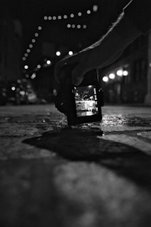 A black and white photograph showing a hand holding a digital camera close to the ground. The camera screen displays a bright street scene with overhead string lights, while the background is blurred. The scene is captured in a dimly lit urban environment.