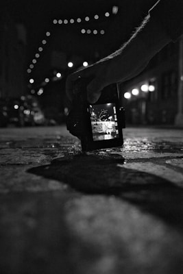 A black and white photograph showing a hand holding a digital camera close to the ground. The camera screen displays a bright street scene with overhead string lights, while the background is blurred. The scene is captured in a dimly lit urban environment.