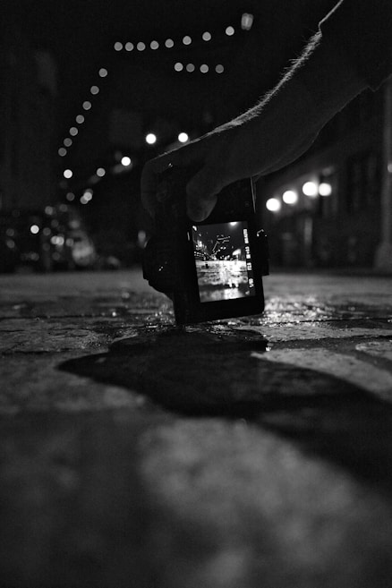A black and white photograph showing a hand holding a digital camera close to the ground. The camera screen displays a bright street scene with overhead string lights, while the background is blurred. The scene is captured in a dimly lit urban environment.