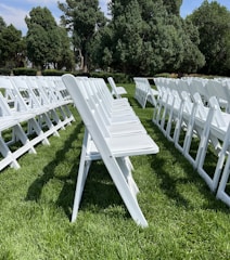 A series of white folding chairs are neatly arranged in rows on a lush green lawn. The scene is framed by tall leafy trees in the background under a clear blue sky. The chairs create an orderly pattern.