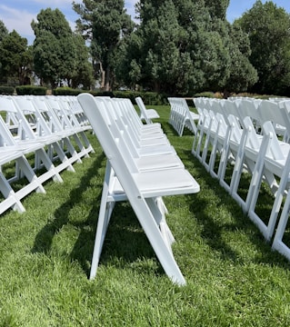 A series of white folding chairs are neatly arranged in rows on a lush green lawn. The scene is framed by tall leafy trees in the background under a clear blue sky. The chairs create an orderly pattern.