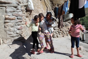 A group of children playfully pose together in front of a rustic stone wall. Two of them hold sticks, while clothing hangs on a line above them. The background is a rural setting with a sunny atmosphere.
