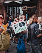 People are gathered in a protest carrying signs and bags. One person prominently holds a sign reading 'Fighting for My Child,' which includes an ultrasound image. The crowd is diverse, with individuals wearing casual clothing and backpacks, standing in an urban setting in front of a storefront.