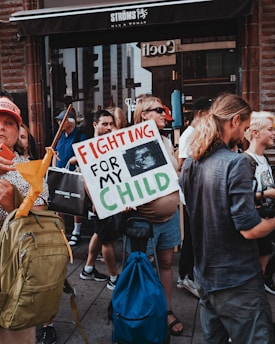 People are gathered in a protest carrying signs and bags. One person prominently holds a sign reading 'Fighting for My Child,' which includes an ultrasound image. The crowd is diverse, with individuals wearing casual clothing and backpacks, standing in an urban setting in front of a storefront.