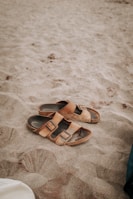 A pair of brown leather sandals lie on a sandy beach, partially covered by sand. The texture of the sand is soft and fine, indicating a warm, leisurely setting. The image captures the simplicity and casual nature of a beach scene.