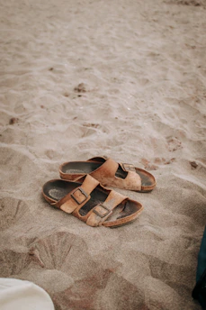 Close-up of brown leather sandals resting on a sandy beach with soft sunlight.