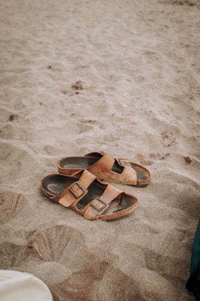 A pair of brown leather sandals lie on a sandy beach, partially covered by sand. The texture of the sand is soft and fine, indicating a warm, leisurely setting. The image captures the simplicity and casual nature of a beach scene.