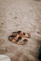 A pair of brown leather sandals lie on a sandy beach, partially covered by sand. The texture of the sand is soft and fine, indicating a warm, leisurely setting. The image captures the simplicity and casual nature of a beach scene.
