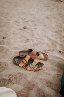 A pair of brown leather sandals lie on a sandy beach, partially covered by sand. The texture of the sand is soft and fine, indicating a warm, leisurely setting. The image captures the simplicity and casual nature of a beach scene.