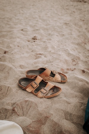 A pair of brown leather sandals lie on a sandy beach, partially covered by sand. The texture of the sand is soft and fine, indicating a warm, leisurely setting. The image captures the simplicity and casual nature of a beach scene.