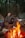 A person sits comfortably in a camping chair, smiling and relaxing by a warm, glowing campfire in the middle of a dense forest. Nearby, a tent and cooler are visible, suggesting a camping trip.