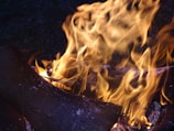 Large orange and yellow flames engulf pieces of dark, charred wood, creating dynamic patterns and intense illumination against a dark background.