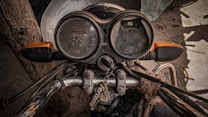 A close-up view of an old motorcycle's dashboard, featuring two circular speedometers, worn and slightly rusty metal parts, and handlebars wrapped in fabric. The worn appearance of the speedometer and the faded brand logo suggest the motorcycle has been in use for several years. The image conveys a sense of nostalgia and ruggedness.