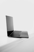 A sleek, open laptop positioned on a white surface with a minimalist design. The screen is tilted back slightly, and the keyboard is visible, showing a modern and streamlined look. The overall setting is bright and clean, accentuating the elegance of the laptop.
