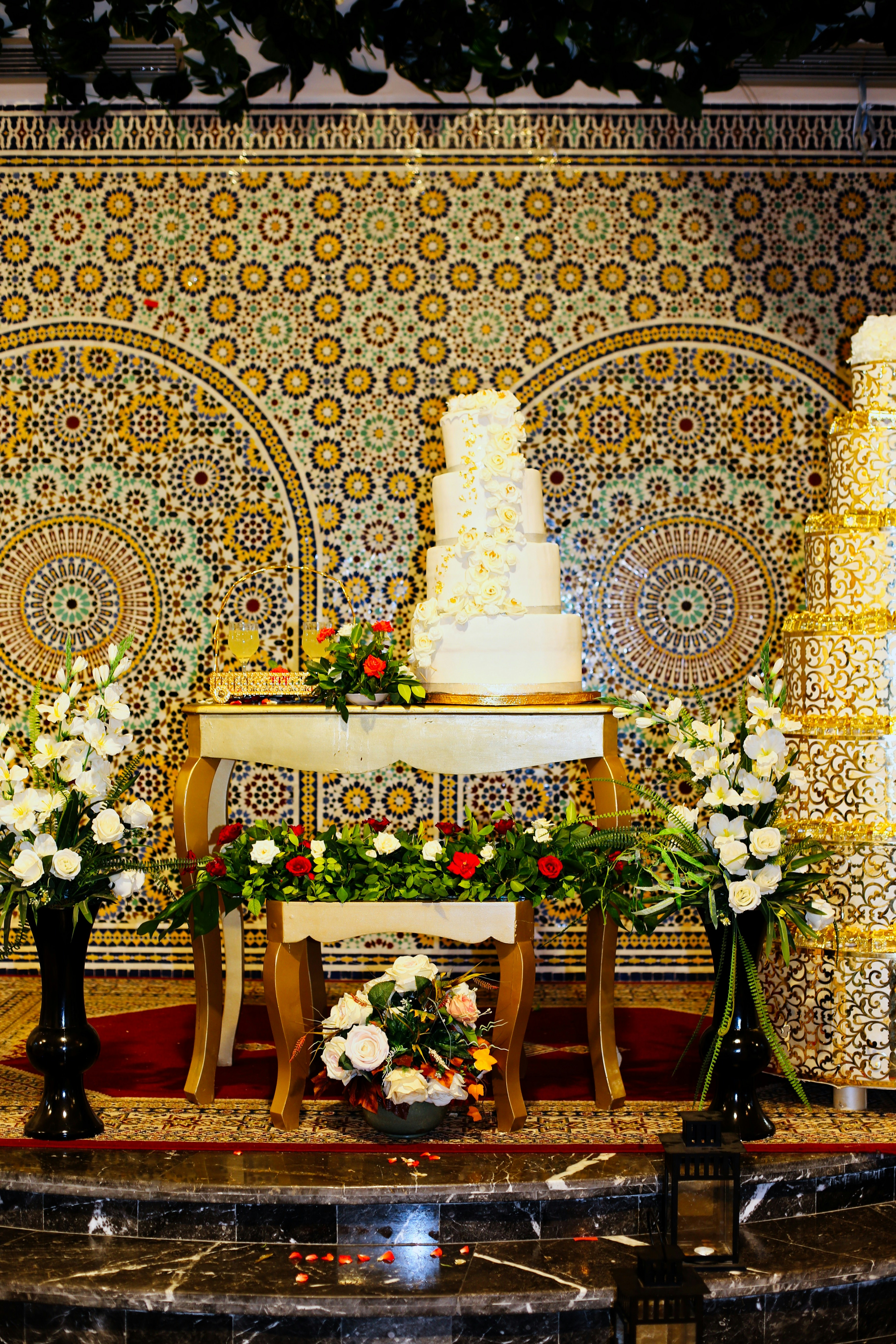 Tall white wedding cake with cascading pink roses and gold ribbon on marble table in sunlit room