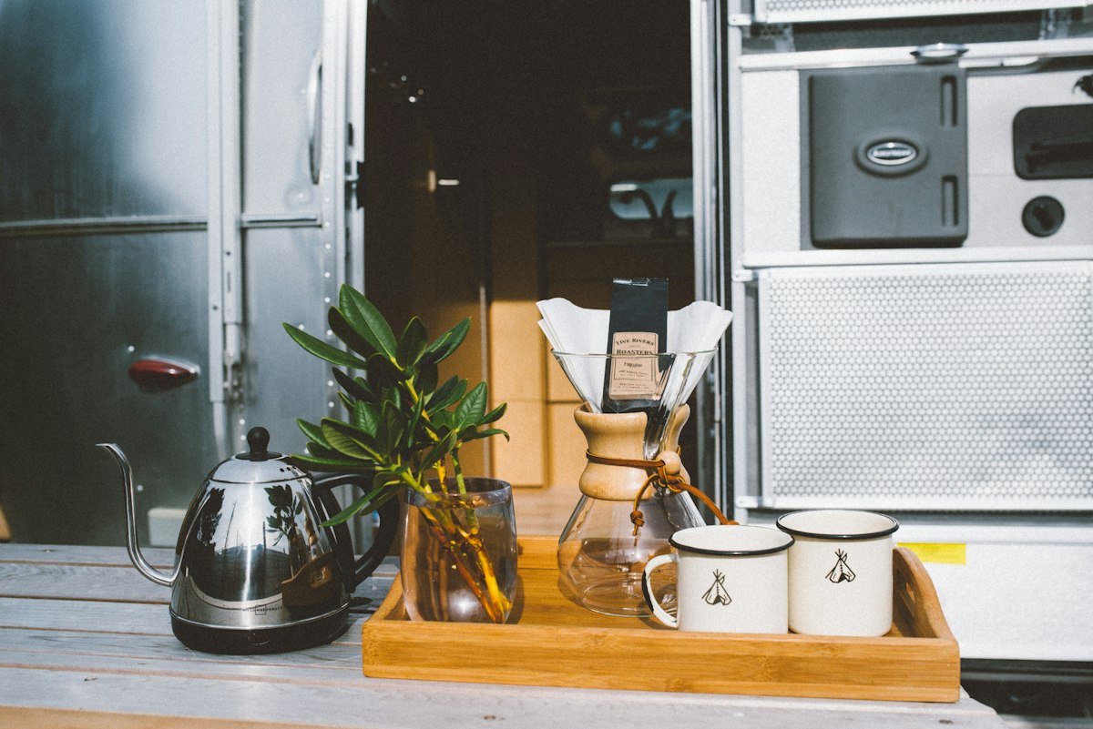 Pava eléctrica de acero con pico cuello de cisne sobre su base, junto a una bandeja de bambú con una cafetera de vidrio y dos tazas enlozadas blancas