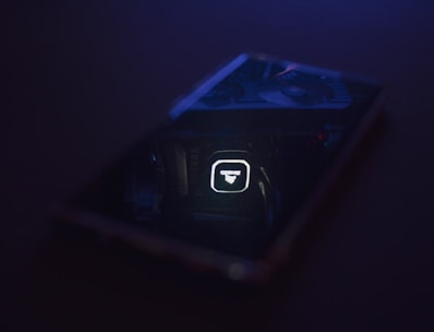 A dimly lit scene featuring a reflective surface, likely a phone or glass, displaying a cooler or fan inside a computer. The reflection shows illuminated text and design on computer hardware.