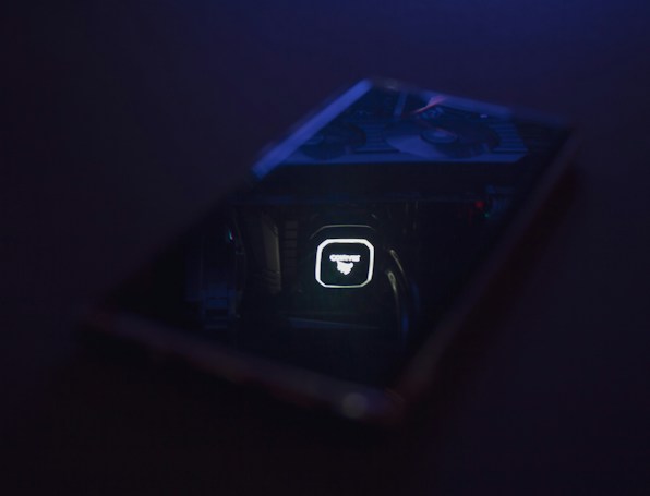 A dimly lit scene featuring a reflective surface, likely a phone or glass, displaying a cooler or fan inside a computer. The reflection shows illuminated text and design on computer hardware.