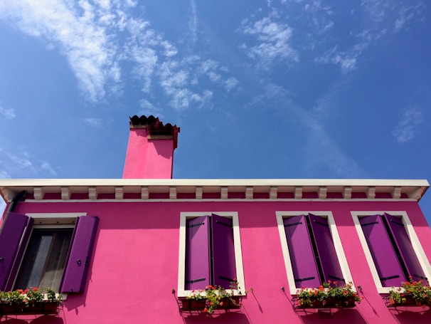 A vibrant pink house with deep purple shutters and a single red chimney stands against a bright blue sky. The facade features several windows adorned with flower boxes containing colorful blooms.