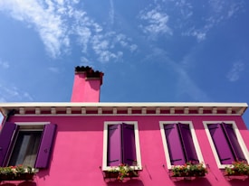 A vibrant pink house with deep purple shutters and a single red chimney stands against a bright blue sky. The facade features several windows adorned with flower boxes containing colorful blooms.