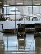 An airplane is parked at a gate, visible through large windows in an airport terminal. Several baggage carts are stationed near a baggage carousel, with tiled floors reflecting the scene. The sky is clear with some buildings in the background.