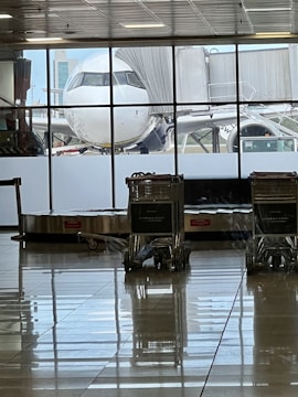 An airplane is parked at a gate, visible through large windows in an airport terminal. Several baggage carts are stationed near a baggage carousel, with tiled floors reflecting the scene. The sky is clear with some buildings in the background.